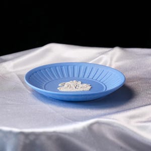 May include: A light blue saucer featuring a white relief design in the center depicting classical figures. The saucer has a shallow bowl shape with a ribbed, textured rim that encircles the central motif, creating a distinct contrast.