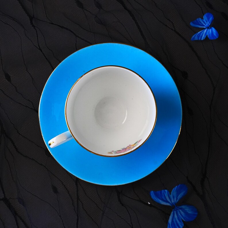 Op de afbeelding: Een felblauwe kop en schotel set met een witte binnenkant en een goudkleurige rand. De kop heeft een klein bloemmotief op de bodem aan de binnenzijde. Twee blauwe vlinderdecoraties bevinden zich bij de schotel op de donkere ondergrond.