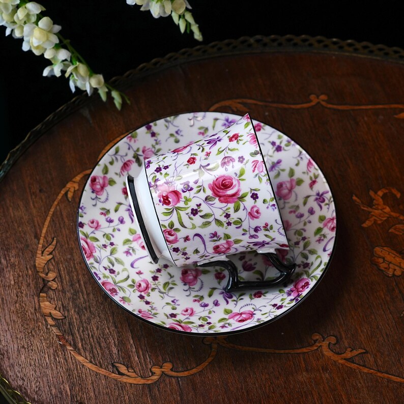 May include: A floral teacup and saucer set featuring pink roses, purple details, and green foliage on a white background. The cup has a black handle and rim, and the saucer has a matching black rim. The set is on a wooden surface.