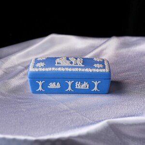May include: A rectangular blue trinket box with white relief detailing. The box features a white floral border and white relief scenes on the lid and sides. The box is set against a white, textured background.