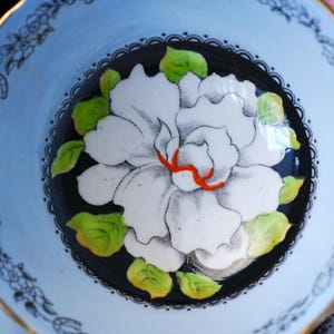 May include: A ceramic bowl featuring a central black circular design with a large white flower, bright green leaves, and orange accents. The outer rim is light blue with a delicate black floral pattern and a thin gold-colored edge.