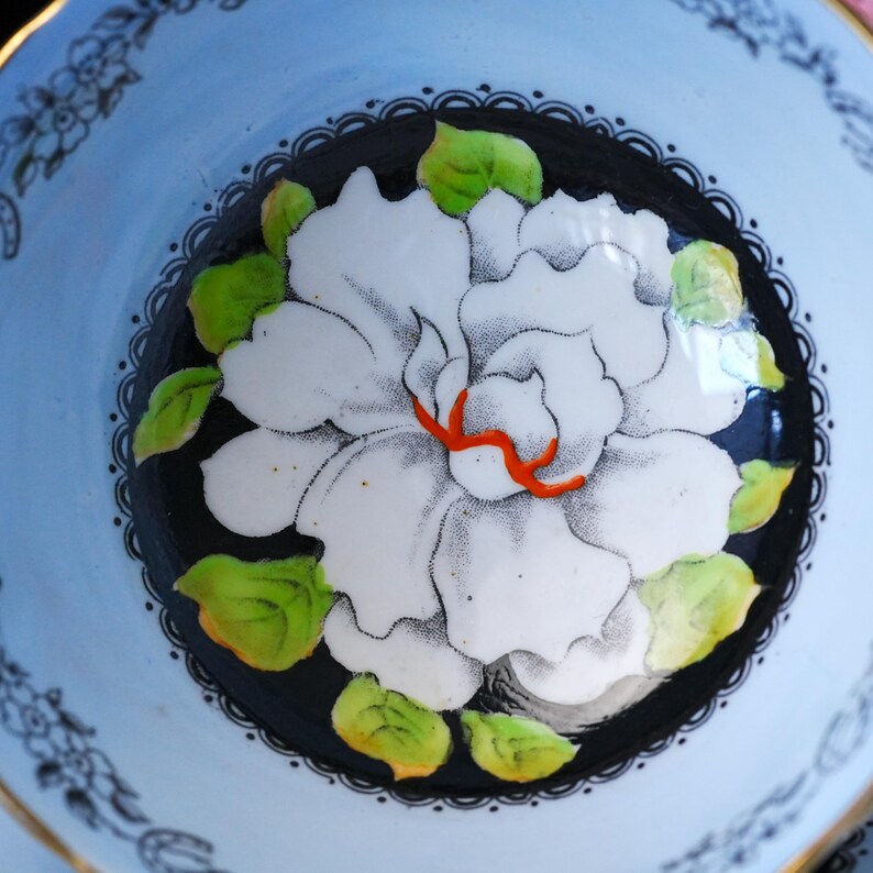 May include: A ceramic bowl featuring a central black circular design with a large white flower, bright green leaves, and orange accents. The outer rim is light blue with a delicate black floral pattern and a thin gold-colored edge.