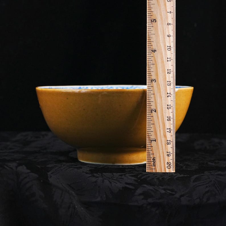 May include: A mustard-yellow ceramic bowl with a blue and white patterned rim. The bowl is approximately 4 inches tall, as measured by a ruler. The bowl is set against a dark background.