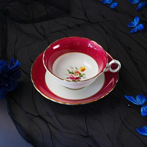 K&ouml;nnte beinhalten: Ein Porzellan-Tassenset mit Untertasse, das einen tief magentafarbenen Rand mit strukturierter Oberfl&auml;che und Goldverzierung aufweist. Die Mitte der Tasse und der Untertassenspiegel sind wei&szlig;, mit einem kleinen, bunten Blumenstrau&szlig; im Tassenboden.