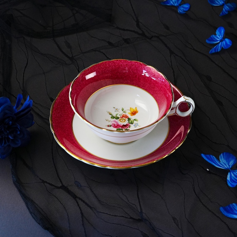 K&ouml;nnte beinhalten: Ein Porzellan-Tassenset mit Untertasse, das einen tief magentafarbenen Rand mit strukturierter Oberfl&auml;che und Goldverzierung aufweist. Die Mitte der Tasse und der Untertassenspiegel sind wei&szlig;, mit einem kleinen, bunten Blumenstrau&szlig; im Tassenboden.