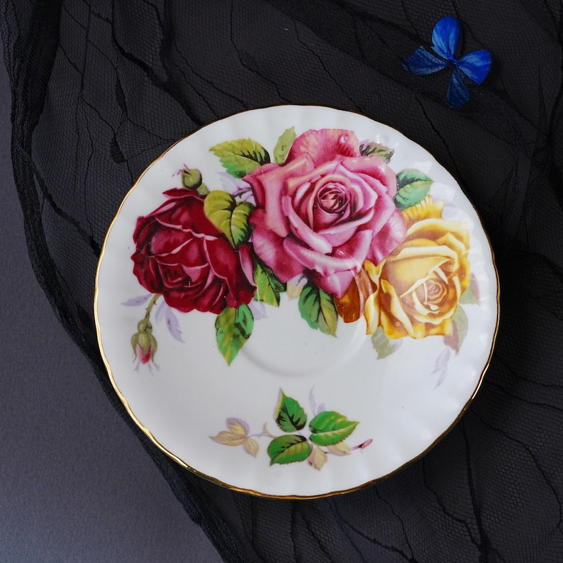 Peut inclure: Une soucoupe en porcelaine blanche avec un bord festonn&eacute; et une bordure dor&eacute;e. Le centre pr&eacute;sente un motif floral avec une rose rouge fonc&eacute;, une grande rose rose et une rose jaune avec des feuilles vertes. Un petit groupe de feuilles est en bas.
