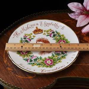 Op de afbeelding: Een wit herdenkingsbord van porselein met een portret van koningin Elizabeth II, een kroon en bloemmotieven. De tekst luidt H.M. Queen Elizabeth II, June 2nd, 1953 en Coronation. Een houten liniaal toont een diameter van 20,3 cm.
