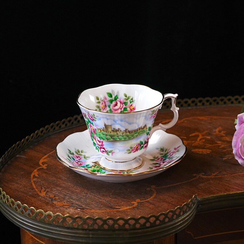 Op de afbeelding: Een witte theekop en schotel met roze rozen en een landschap. De kop en schotel hebben een gouden rand. Het landschap toont een kasteel. De set staat op een houten tafel.