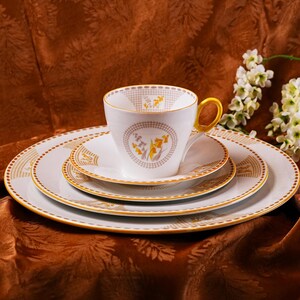 Op de afbeelding: Een witte theekop met een gouden oor en bijpassende schotels en borden. De theekop en borden hebben een gouden en gele bloemenprint. De theekop en borden staan op een bruin, patroon tafelkleed. Een takje witte bloemen staat op de achtergrond.