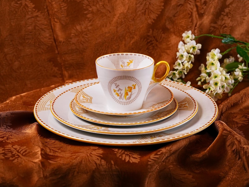 Op de afbeelding: Een witte theekop met een gouden oor en bijpassende schotels en borden. De theekop en borden hebben een gouden en gele bloemenprint. De theekop en borden staan op een bruin, patroon tafelkleed. Een takje witte bloemen staat op de achtergrond.