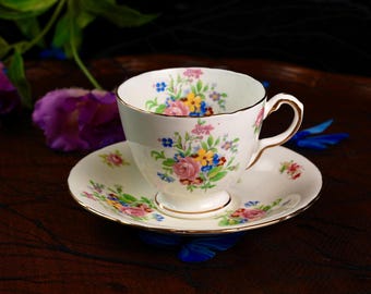 May include: A white teacup and saucer set with a floral pattern in pink, yellow, blue, and green. The teacup has a gold rim and handle. The saucer also has a matching floral design. A classic tea set.