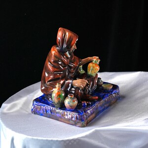 May include: A ceramic figurine of a robed figure seated on a blue base, with small, colourful vases. The figure wears a brown hooded cloak. The rectangular base has a textured, glazed finish. The vases have orange, green, and blue accents.