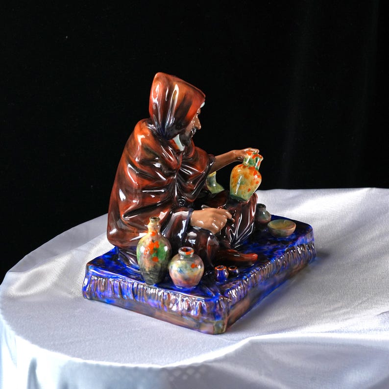 May include: A ceramic figurine of a robed figure seated on a blue base, with small, colourful vases. The figure wears a brown hooded cloak. The rectangular base has a textured, glazed finish. The vases have orange, green, and blue accents.