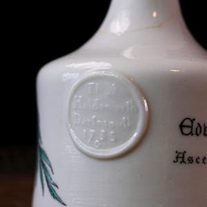 K&ouml;nnte beinhalten: Nahaufnahme einer wei&szlig;en Keramikflasche mit abgerundeter Schulter und schmalem Hals. Ein kreisf&ouml;rmiges gepr&auml;gtes Siegel mit Text ist sichtbar, zusammen mit schwarzer Schrift und einer gr&uuml;nen botanischen Illustration. Die Flasche hat eine Vintage-&Auml;sthetik.