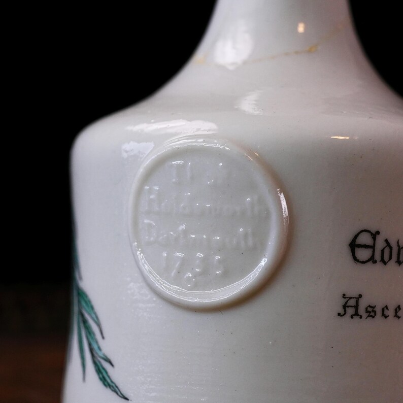 K&ouml;nnte beinhalten: Nahaufnahme einer wei&szlig;en Keramikflasche mit abgerundeter Schulter und schmalem Hals. Ein kreisf&ouml;rmiges gepr&auml;gtes Siegel mit Text ist sichtbar, zusammen mit schwarzer Schrift und einer gr&uuml;nen botanischen Illustration. Die Flasche hat eine Vintage-&Auml;sthetik.