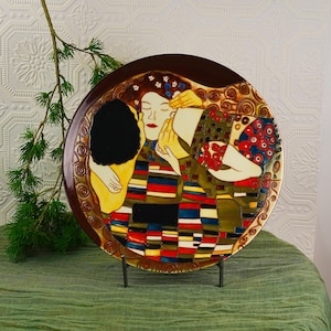 May include: A decorative ceramic plate featuring a colorful, abstract design with figures embracing. The plate has a brown rim and is displayed on a black metal stand. The artwork includes red, blue, and yellow hues.