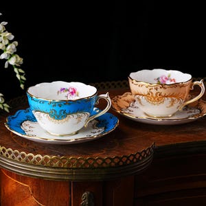 Op de afbeelding: Twee sierlijke kopjes en schotels. E&eacute;n kopje is turquoise met gouden accenten, de andere perzik met gouden filigraan. Beide hebben bloemmotieven en staan op een houten tafel. Witte bloemen zijn op de achtergrond.