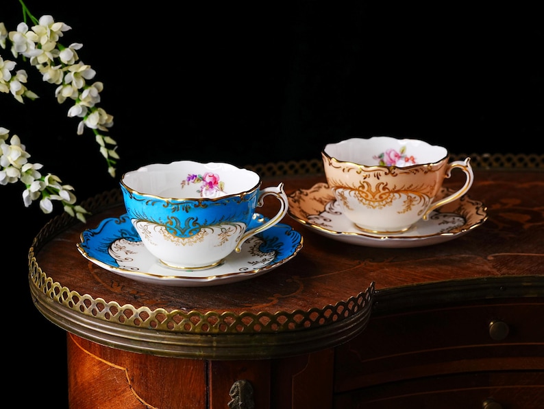 Op de afbeelding: Twee sierlijke kopjes en schotels. E&eacute;n kopje is turquoise met gouden accenten, de andere perzik met gouden filigraan. Beide hebben bloemmotieven en staan op een houten tafel. Witte bloemen zijn op de achtergrond.