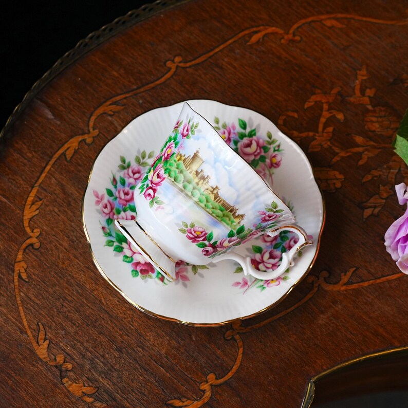 Op de afbeelding: Een witte theekop en schotel set met een bloemenpatroon van roze rozen en groene bladeren. De theekop heeft een landschapssc&egrave;ne en gouden rand. De set staat op een houten tafel.