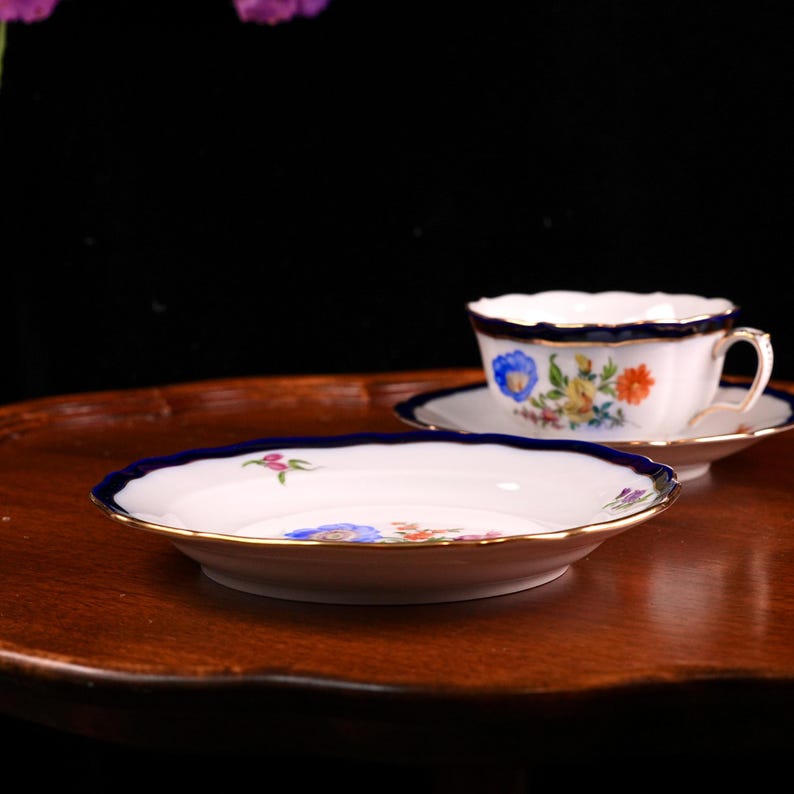 Puede incluir: Un juego de taza y platillo blancos con borde azul y ribete dorado. La taza y el platillo presentan un dise&ntilde;o floral con flores azules, amarillas y naranjas. El juego est&aacute; sobre una superficie de madera.