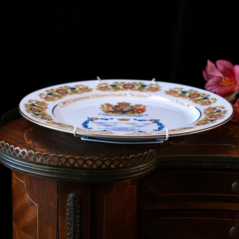 Pode incluir: Um prato comemorativo de cer&acirc;mica branca com um bras&atilde;o real ao centro, uma borda dourada e uma moldura decorativa com motivos florais. O texto diz: To Commemorate H.M. Queen Elizabeth The Queen Mother's 90th Birthday 1990.
