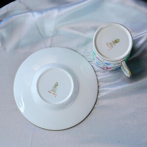 May include: A white porcelain teacup and saucer set featuring a delicate floral pattern with pink and blue flowers. The underside of both pieces displays a gold maker's mark and the red text WN 1017, indicating the origin as made in England.