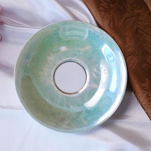 May include: A vintage iridescent saucer with a central white circle and a gold rim. The saucer has a pearlescent finish with shades of green and blue. The saucer is likely part of a tea set or a decorative item.