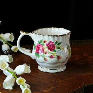 Peut inclure: Une tasse &agrave; th&eacute; en porcelaine blanche avec un bord festonn&eacute; dor&eacute;, une anse incurv&eacute;e aux accents dor&eacute;s et un motif floral de roses roses avec des feuilles vertes. La tasse pr&eacute;sente un corps cannel&eacute; et une base sur pied avec un bord dor&eacute;.