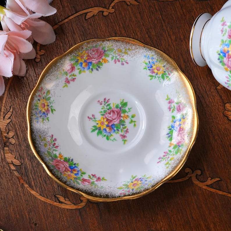 Może przedstawiać: Biały porcelanowy spodek ze złotym, falistym brzegiem, ozdobiony kwiatowymi bukietami w kolorze r&oacute;żowym, ż&oacute;łtym, niebieskim i zielonym. Środek zdobi bukiet r&oacute;ż. Częściowo widoczny pasujący filiżanka.