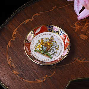 May include: A round porcelain commemorative dish featuring the Royal Coat of Arms of the United Kingdom in the center. The text reads QUEEN ELIZABETH II GOLDEN JUBILEE 1952-2002. The border is decorated with red, blue, and gold floral patterns.
