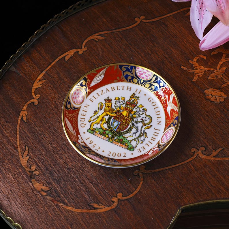 May include: A round porcelain commemorative dish featuring the Royal Coat of Arms of the United Kingdom in the center. The text reads QUEEN ELIZABETH II GOLDEN JUBILEE 1952-2002. The border is decorated with red, blue, and gold floral patterns.