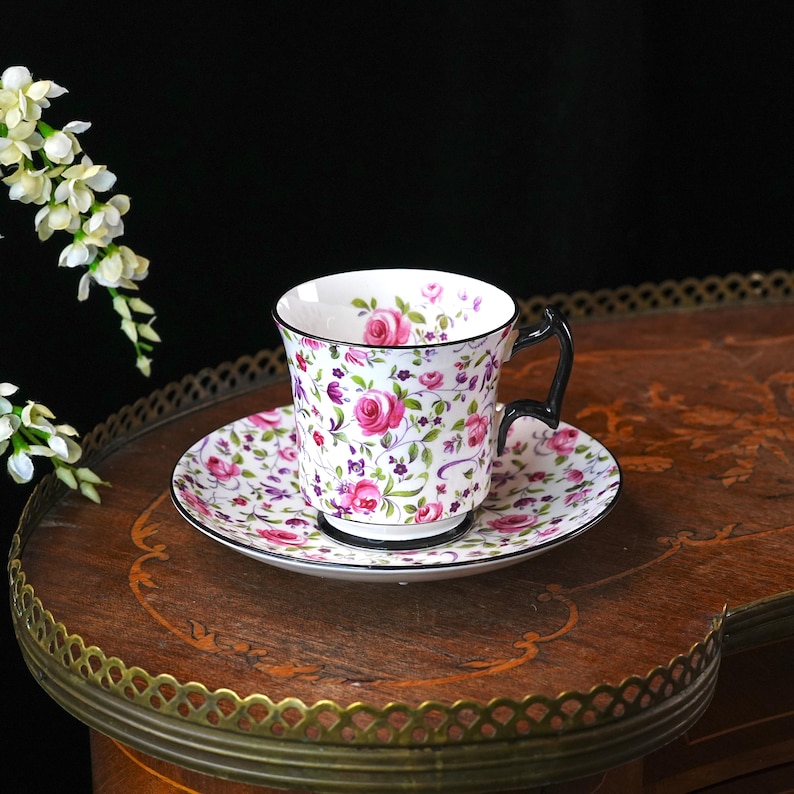 May include: A floral teacup and saucer set. The white porcelain cup and saucer feature a pattern of pink roses, purple accents, and green leaves. The cup has a black handle and trim. The set is on a wooden table.
