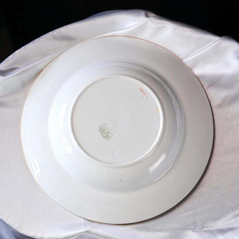 May include: A white porcelain plate with a gold rim, viewed from the underside. The plate features a central recessed area and a wider outer rim. A small, faded emblem and text are visible near the center. The plate is set against a white fabric background.