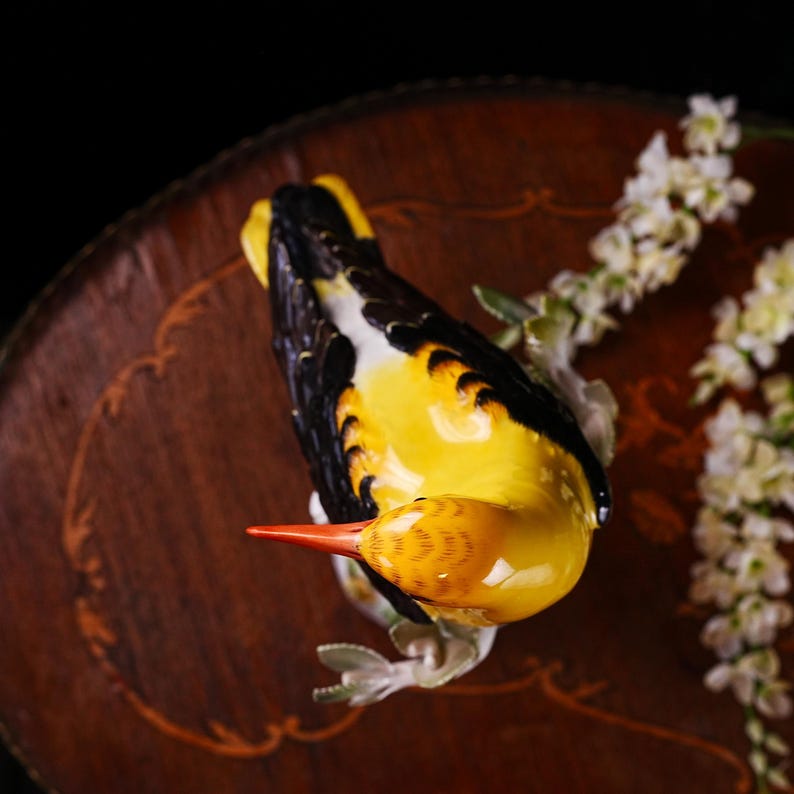 May include: A ceramic bird figurine with yellow, black, and white plumage, perched on a branch with white flowers. The bird has an orange beak and is set against a dark background.