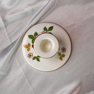 Op de afbeelding: Een witte keramische kandelaar met een gouden rand en een aardbeienontwerp. Het ontwerp bevat groene bladeren, roze bloemen en rode bessen. De kandelaar is rond en heeft een kleine, centrale beker.