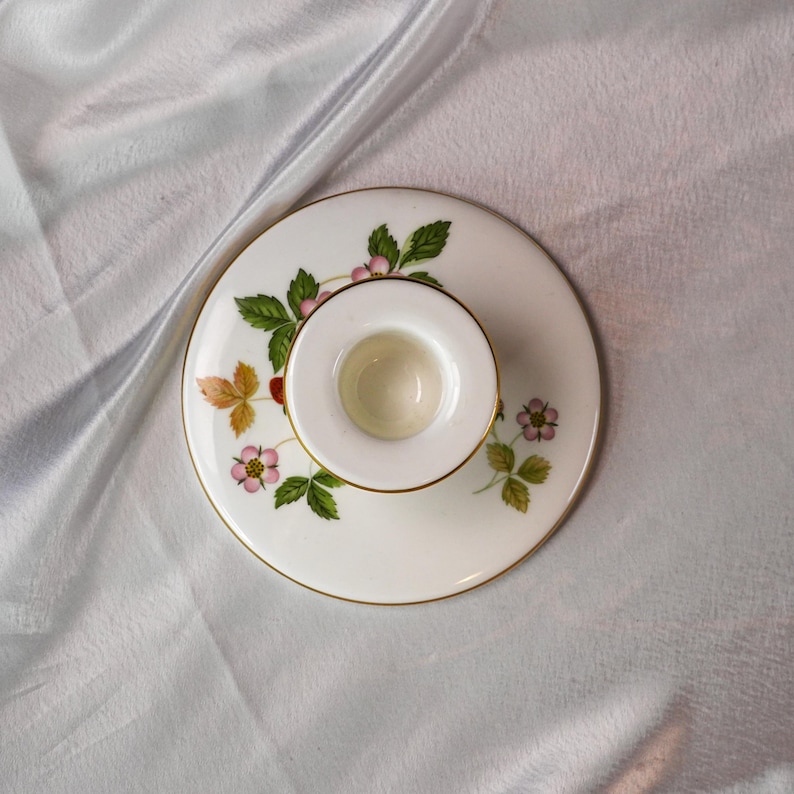 Op de afbeelding: Een witte keramische kandelaar met een gouden rand en een aardbeienontwerp. Het ontwerp bevat groene bladeren, roze bloemen en rode bessen. De kandelaar is rond en heeft een kleine, centrale beker.
