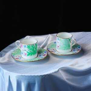 May include: Two white porcelain cups and matching saucers featuring a hand-painted botanical pattern of vibrant green leaves and small pink and blue flowers. Each piece is finished with a thin gold rim along the top edge and saucer border.
