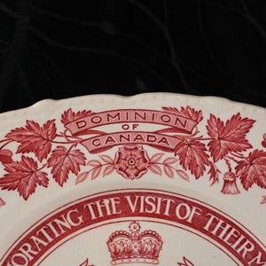 May include: A cream-coloured ceramic plate featuring a red transferware design. The text reads DOMINION OF CANADA and COMMEMORATING THE VISIT OF THEIR M. A central crown icon is surrounded by maple leaves and floral motifs with a scalloped edge.