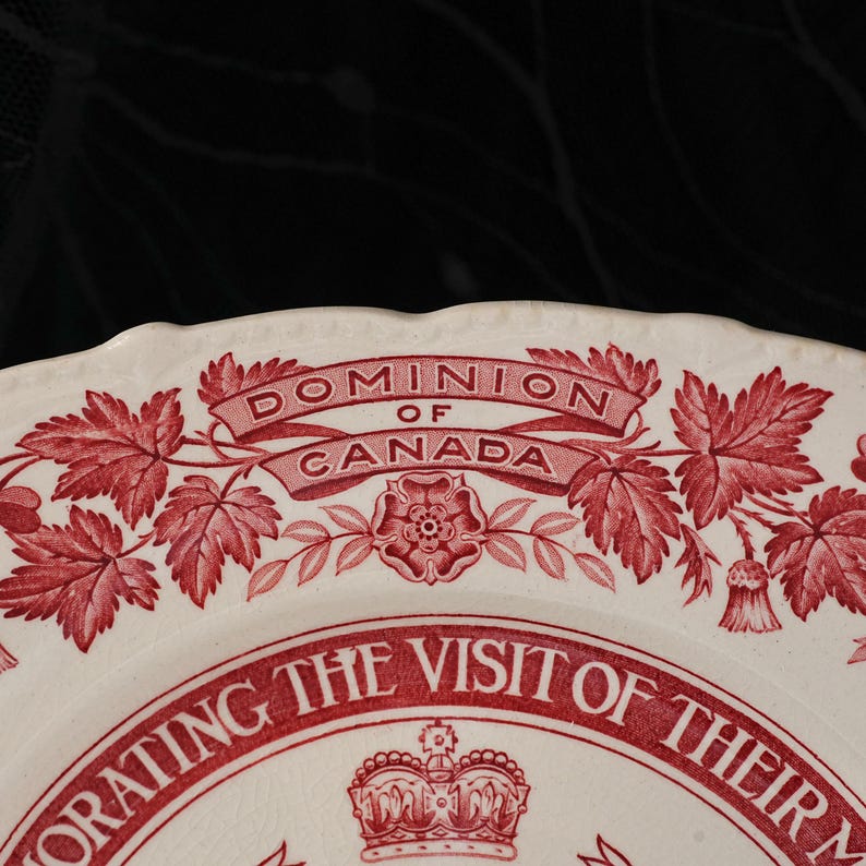 May include: A cream-coloured ceramic plate featuring a red transferware design. The text reads DOMINION OF CANADA and COMMEMORATING THE VISIT OF THEIR M. A central crown icon is surrounded by maple leaves and floral motifs with a scalloped edge.