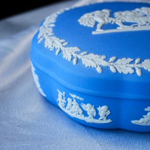 May include: A round blue ceramic trinket box featuring white relief ornamentation. The lid displays a detailed classical scene encircled by a leafy garland, while the side of the base is decorated with a continuous frieze of small figures.