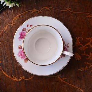 Op de afbeelding: Een witte theekop en schotelset met een gouden rand en roze rozenbloemmotief. De theekop staat op de schotel en toont de fijne details van de vintage theeset. De set staat op een donkere houten ondergrond.