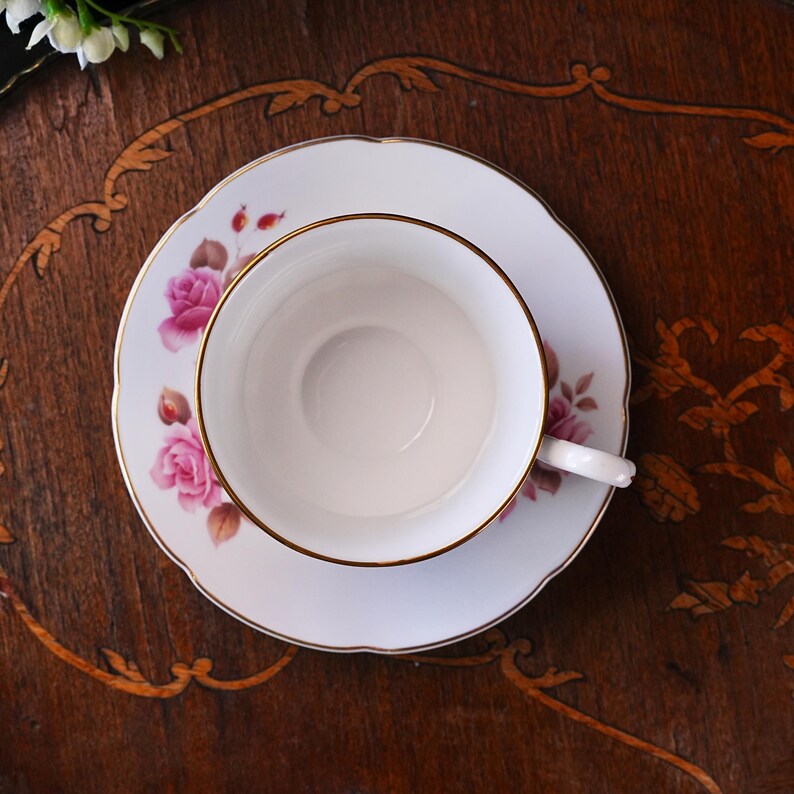 Op de afbeelding: Een witte theekop en schotelset met een gouden rand en roze rozenbloemmotief. De theekop staat op de schotel en toont de fijne details van de vintage theeset. De set staat op een donkere houten ondergrond.