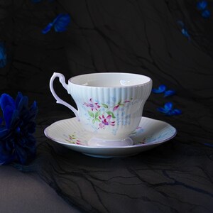 May include: A white porcelain teacup and saucer set featuring a ribbed texture and a delicate floral pattern of pink fuchsia blossoms and small blue sprigs. The cup has a curved handle and a flared rim, while the saucer has a matching scalloped edge.