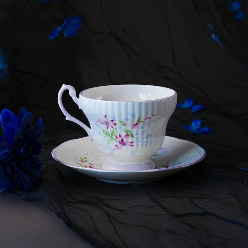 May include: A white porcelain teacup and saucer set featuring a ribbed texture and a delicate floral pattern of pink fuchsia blossoms and small blue sprigs. The cup has a curved handle and a flared rim, while the saucer has a matching scalloped edge.