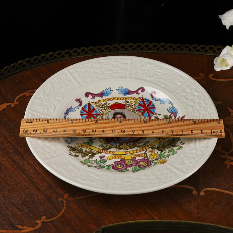 Op de afbeelding: Een wit keramieken herdenkingsbord met een portret van koningin Elizabeth II, de tekst 40th ANNIVERSARY OF HM QUEEN ELIZABETH 1952-1992 en bloemmotieven. Een houten liniaal op het bord toont een diameter van ongeveer 20 cm.