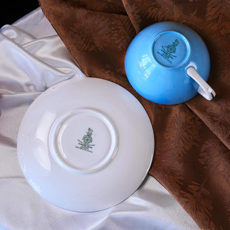 Pode incluir: Um conjunto de ch&aacute;vena e pires azul claro. A ch&aacute;vena &eacute; azul celeste com uma pega branca. O pires &eacute; branco com um emblema verde no centro. O conjunto est&aacute; sobre uma superf&iacute;cie com padr&atilde;o castanho e branco.