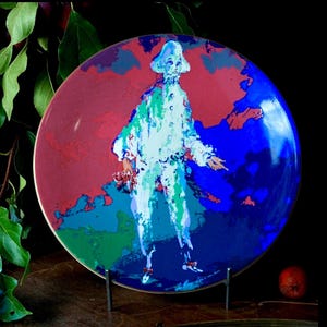 May include: A decorative plate with a stylized figure in a white costume with blue and green accents, holding a small bouquet. The background is a mix of red and blue, with a dark border. The plate is displayed on a metal stand.