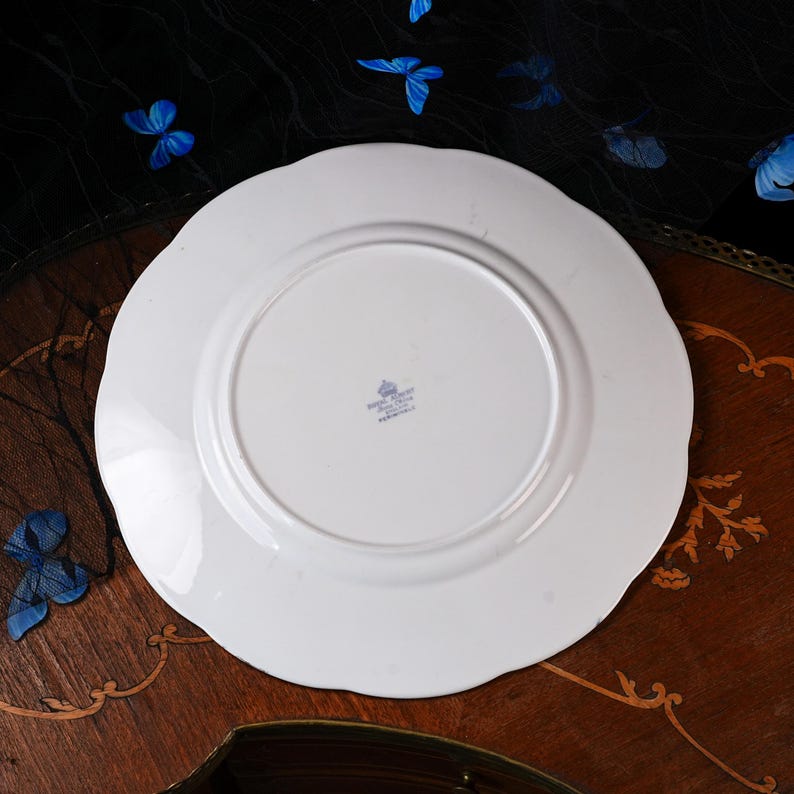 Puede incluir: Un plato de cer&aacute;mica blanca con borde festoneado. La parte inferior presenta una marca de fabricante impresa en azul que dice Royal Albert Bone China England. El plato tiene un acabado liso y brillante y un anillo de base circular central.