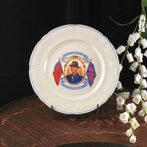 May include: A cream-colored ceramic commemorative plate featuring a central portrait of King Edward VIII in a military uniform, flanked by two flags. The text reads H.M. KING EDWARD VIII and ACCESSION 21.1.1936, with a blue rim and embossed edge.