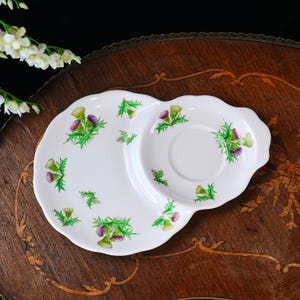 Puede incluir: Plato de cer&aacute;mica blanca con un platillo incorporado, decorado con hojas verdes y flores de cardo moradas. El plato tiene un borde festoneado y est&aacute; colocado sobre una superficie de madera oscura. Un dise&ntilde;o floral est&aacute; pintado en el plato.